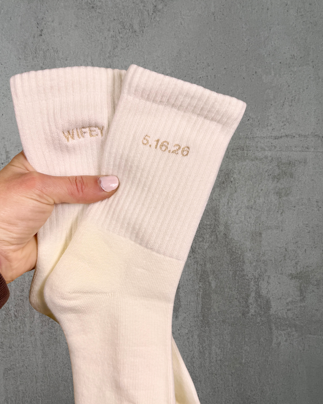 a pair of white socks with the text "wifey" and a wedding date is held up against a gray background