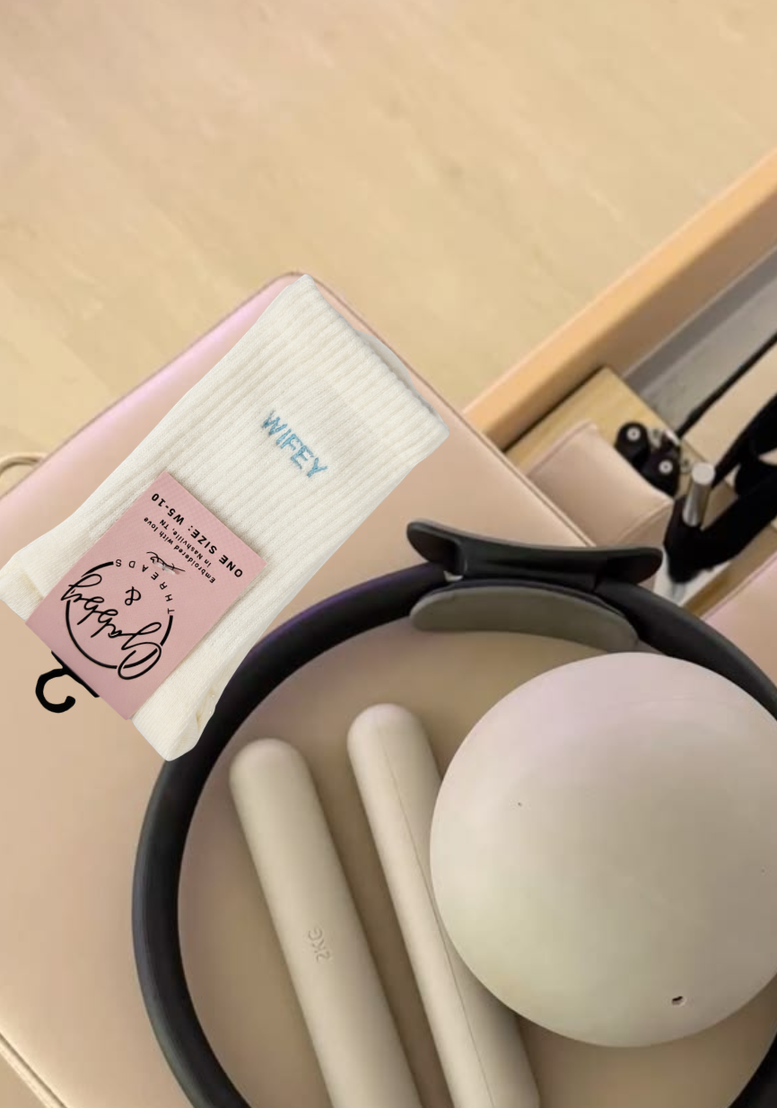 a pair of white grip socks with the word "wifey" embroidered on them sits next to workout equipment on a reformer