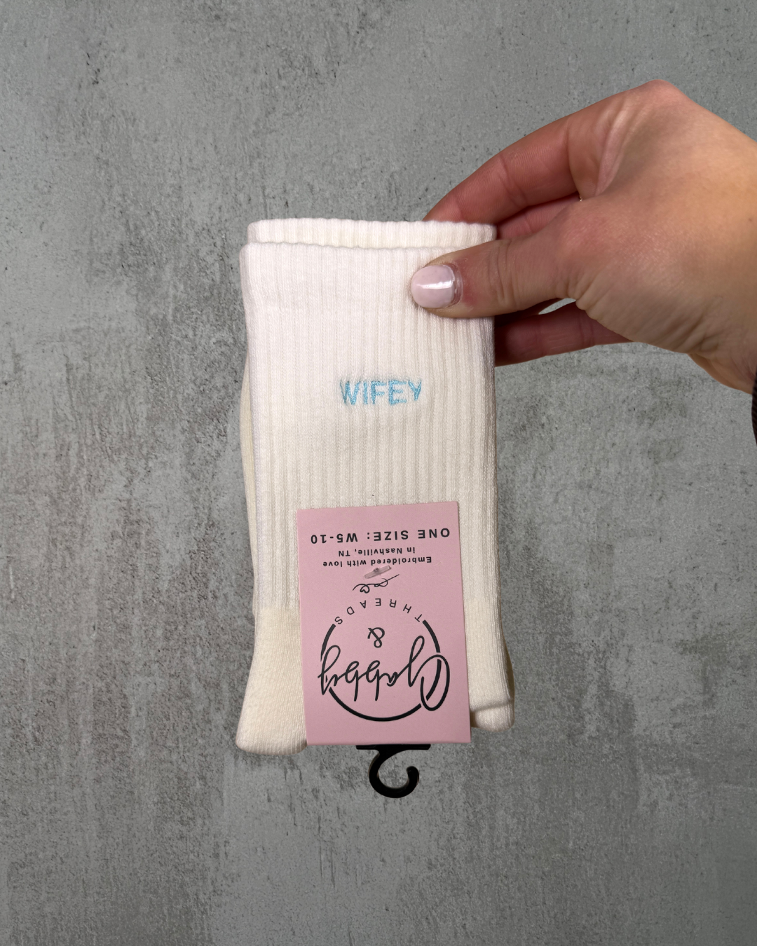 White grip sock with 'WIFEY' text held by a hand on a gray tiled floor.
