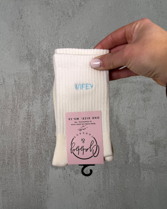 White grip sock with 'WIFEY' text held by a hand on a gray tiled floor.
