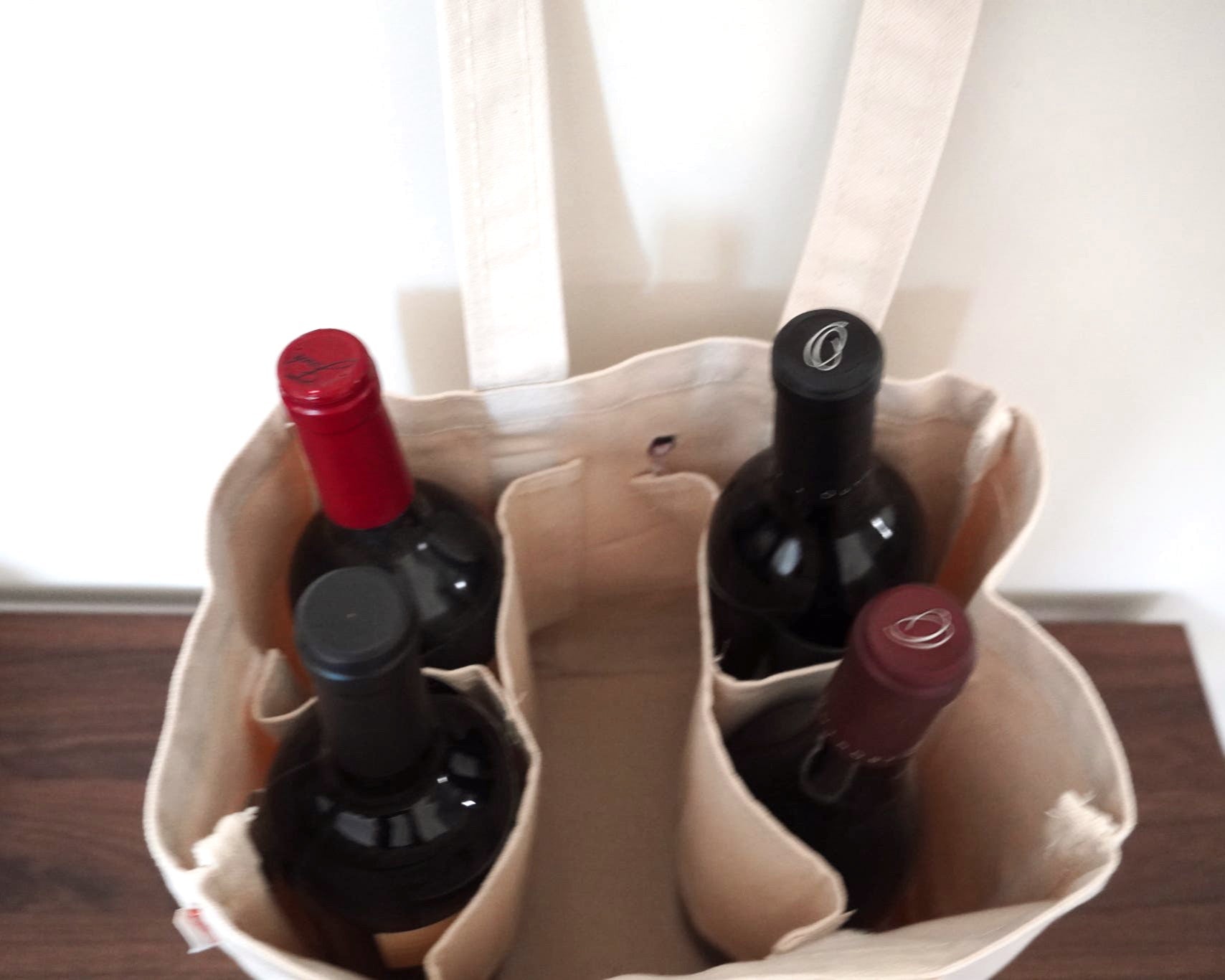 Four wine bottles in a beige tote bag on a wooden surface with a blurred background