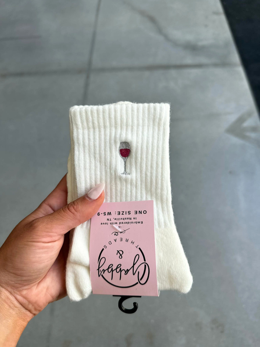 a pair of white grip socks with an embroidered wine glass on them held up by a hand against a grey background