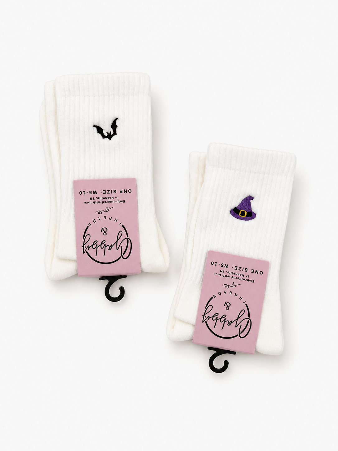 Two pairs of white crew socks on black hangers against a white background. One pair features a small black bat embroidery, and the other features a small purple witch hat embroidery.
