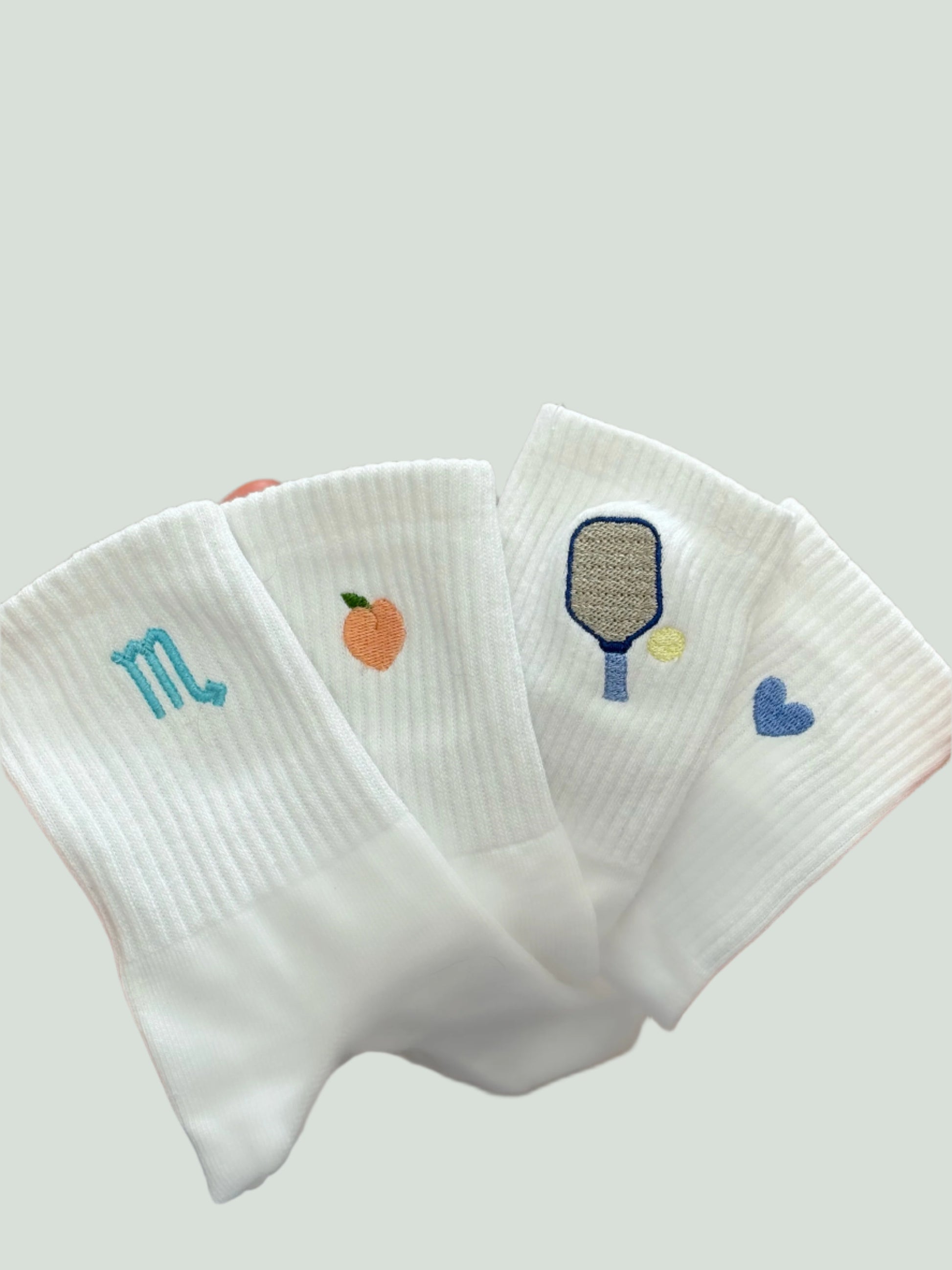 An image with four pairs of socks all featuring different embroidered designs (zodiac symbol, peach, pickleball paddle and heart)